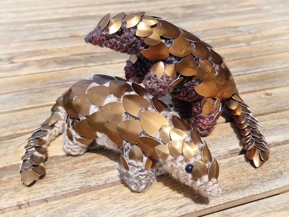 Two knitted pangolins with bronze scales, one with light brown skin standing on a wooden table, and one with dark brown skin beside it with front legs climbing onto the light brown one.
